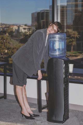 Tired woman in grey suit leaning her head on the water cooler in an office