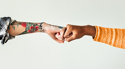 Arms from two people fist bumping. One arm is heavily tattooed the other arm has an orange sleeve.