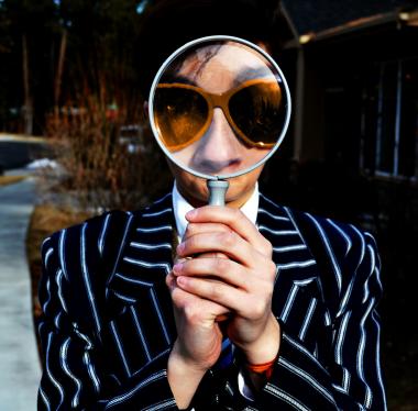 Person in pinstripe suit and orange sunglasses holding a magnifying glass up to his face.